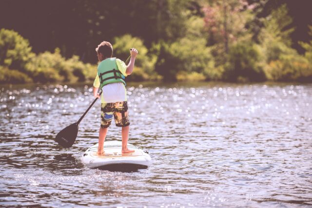 A young boy in a green life vest, shown from behind, stand-up paddle boards on a lake. Kid-friendly fun at Lake Natoma involves stand-up paddle boarding and kayaking