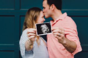 An expectant couple stand together as they hold up an ultrasound photo of their baby. The man is kissing the forehead of his pregnant wife.