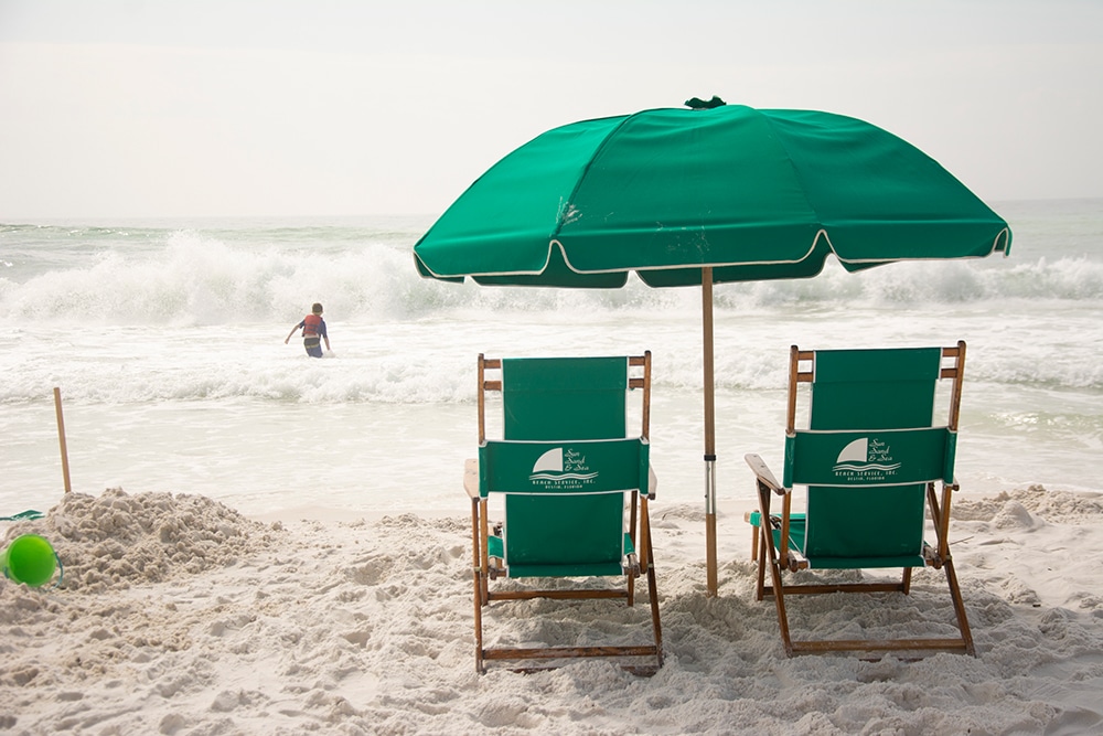Green beach chairs and a matching umbrella with the ocean in the distance. A child is playing in the waves. One last beach visit is a great recipe for Labor Day fun. 