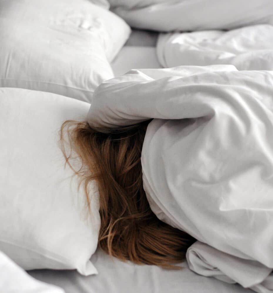 A person is asleep in bed with only their hair peeking out from the white covers. Travel hacks for tired moms.