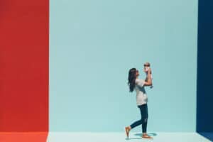 Travel hacks for Tired Moms. A woman in a white t-shirt and jeans holds up her baby while she walks in front of a colorful wall.