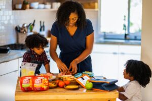 A woman prepares a sandwich as two children stand on either side of her and watch. Managing food allergies while on vacation