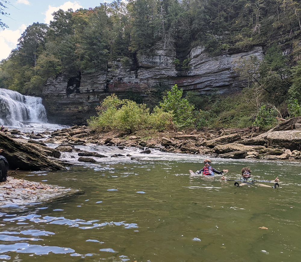 Kids wave at the camera while swimming near a waterfall. Our first camping trip.