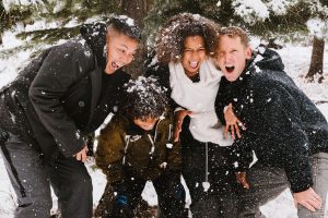A family laughs as they huddle in the cold. Snow falls around them and covers their heads in a white dusting.