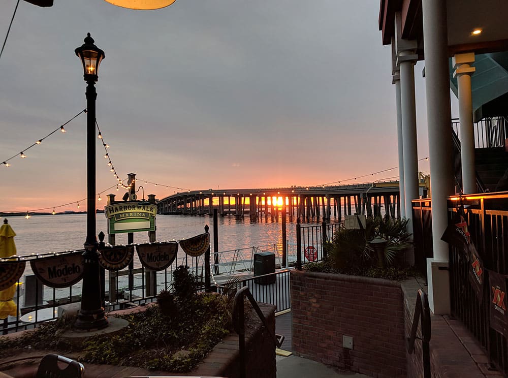 The sun sets over Destin Harbor on the Gulf Mexico, with a long bridge in the background. HarborWalk Village has a beautiful view and is a great place for families.
