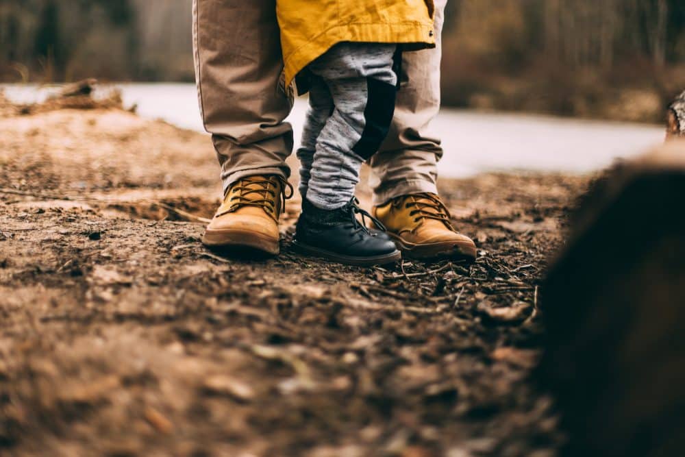 A dad stands with a young child, only visible from the knees down. They are wearing hiking boots and are clearly out for adventures.