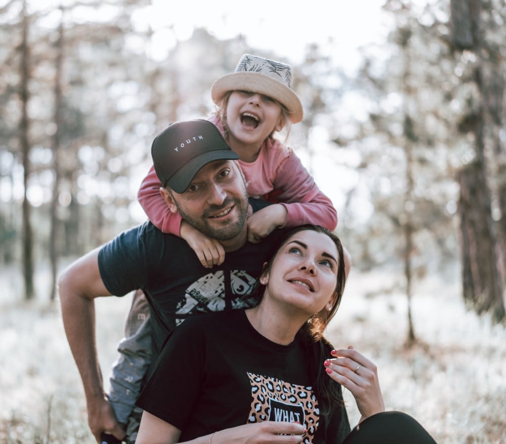 A little girl climbs on top of her two parents while they are hiking in the woods. 