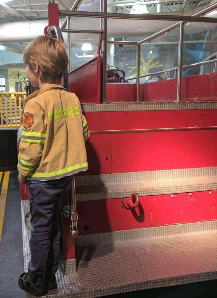 A young boy dressed as a firefighter stands on a fire truck inside a children's museum. 