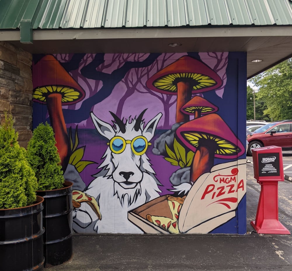 A purple-tinged mural of a cartoon goat wearing sunglasses and holding a box of pizza. Mountain Goat Market in Monteagle, TN.