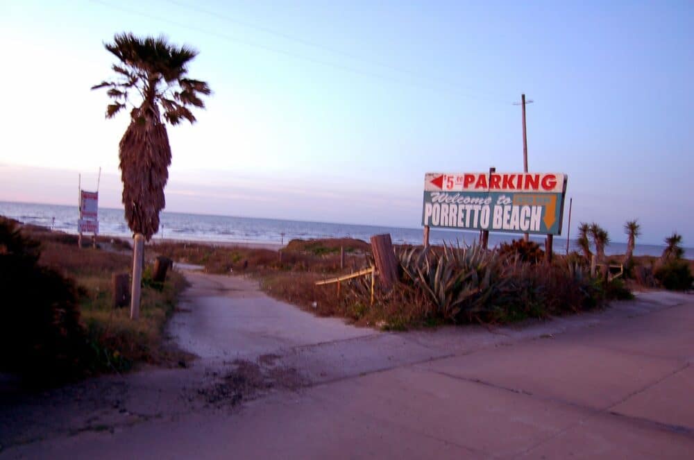 A small gravel footpath branches off from a sidewalk. A sign indicates parking for Porretto Beach. A small palm tree is off to the left of the path.