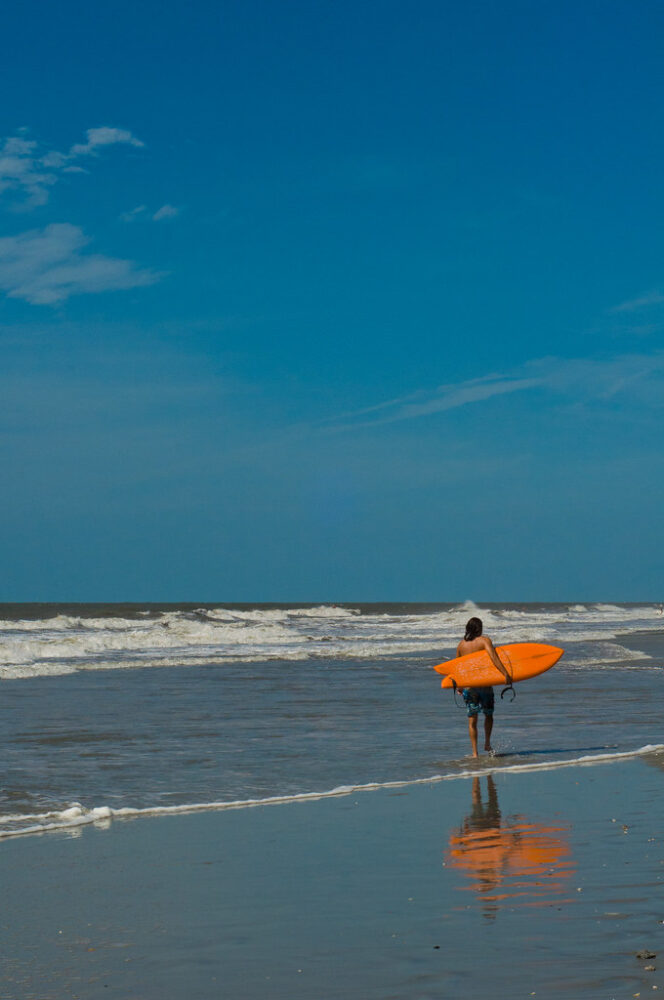 Man carrying surfboard on Folly Beach - 5 Best Charleston Beaches for Epic Family Vacations