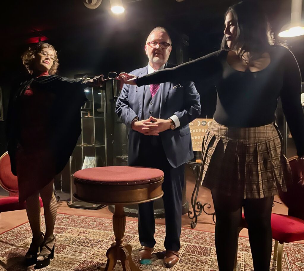 two women touch fingers across table with psychic man in suit behind them
