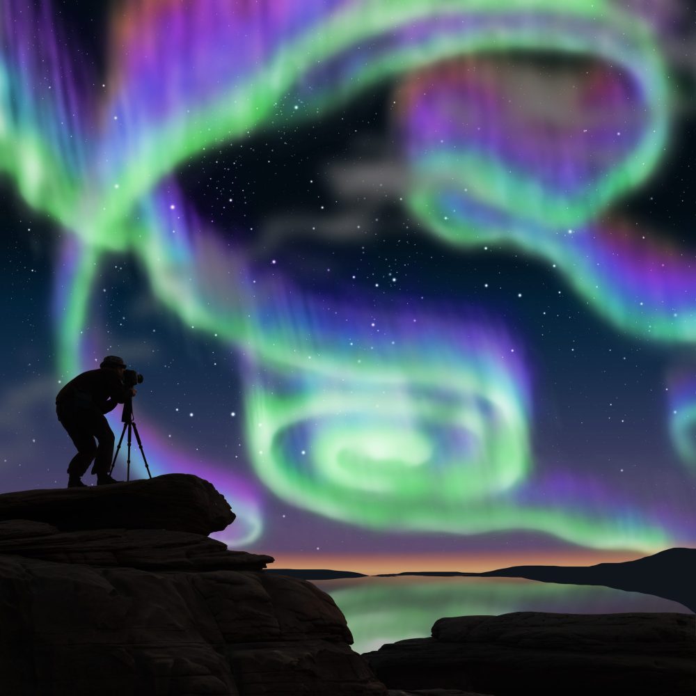 photographer stands atop a mountain photographing the swirling northern lights