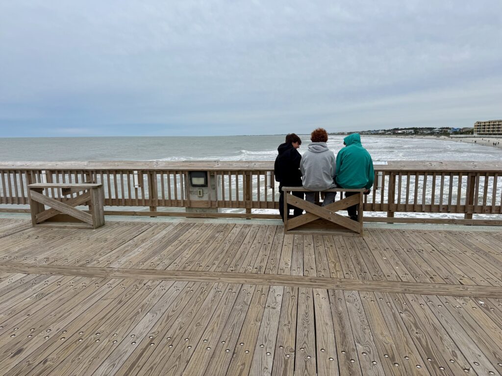 three boys sitting on folly pier looking at ocean