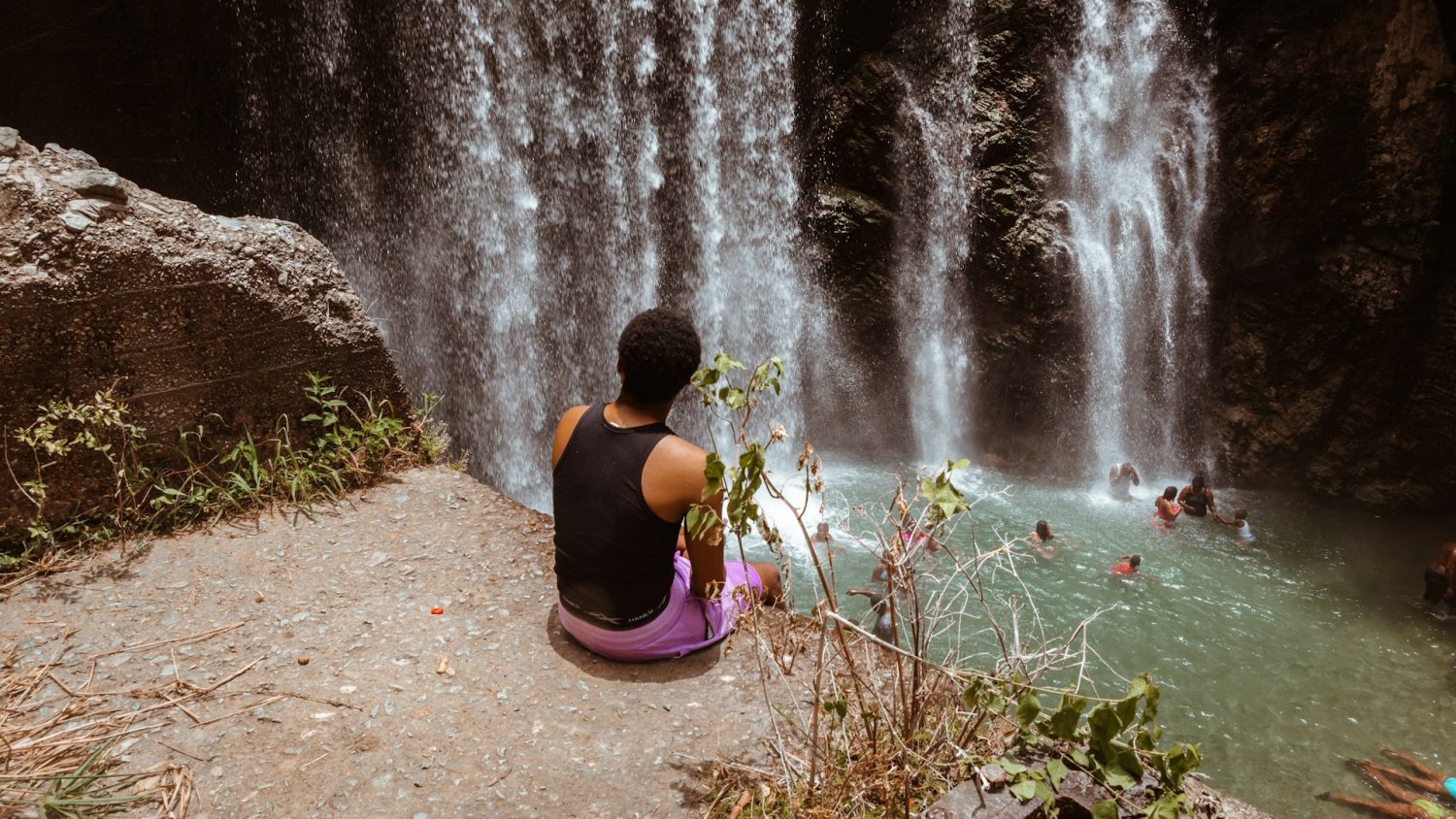 Boy sitting on ledge of waterfall with a family swimming in the pool down below. Best hotels in Jamaica for families.