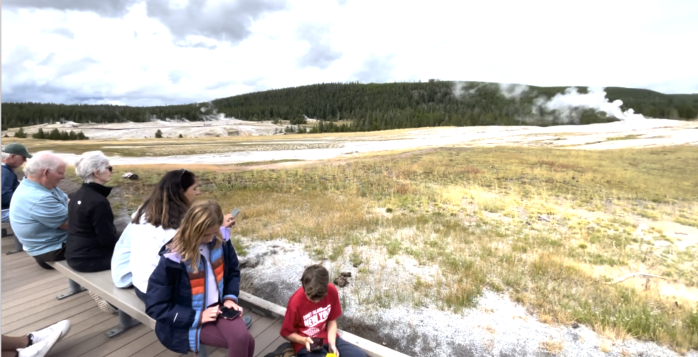 families sitting on benches and boardwalk looking at steam of Old Faithful coming out of ground