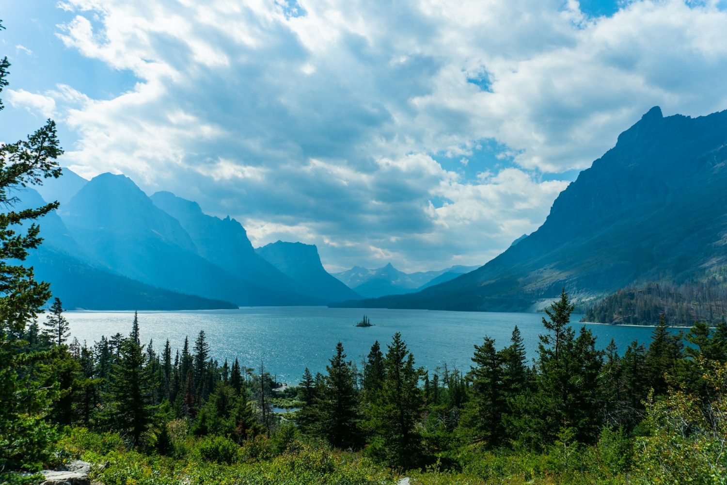 View of mountain range and fir trees surrounding a lake. Things to do in Glacier National Park Montana. 