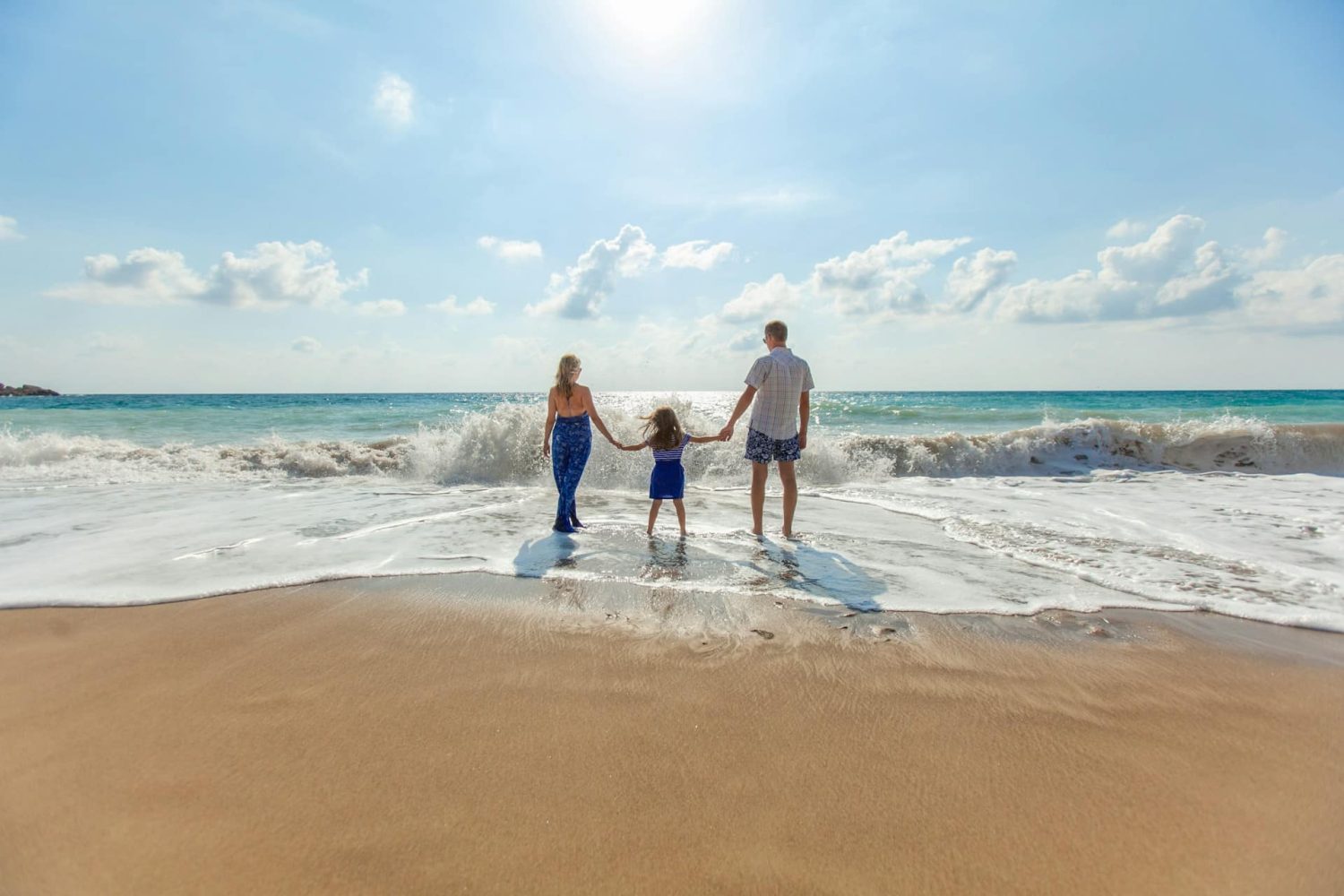 Parents holding hands with child, waiting for waves to splash up on the beach.