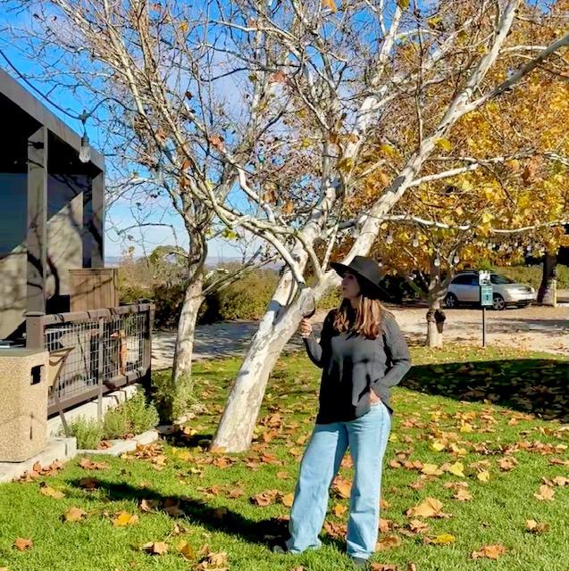 Woman outdoors enjoying a glass of wine during a road trip through California’s Central Coast, with famous winery and trees in the background.