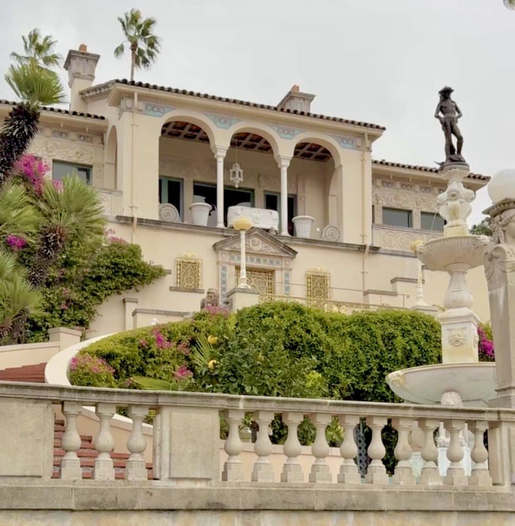 Wide view of Hearst Castle perched atop the hills, showcasing its grand architecture, turrets, and sprawling estate with lush greenery surrounding the historic mansion.