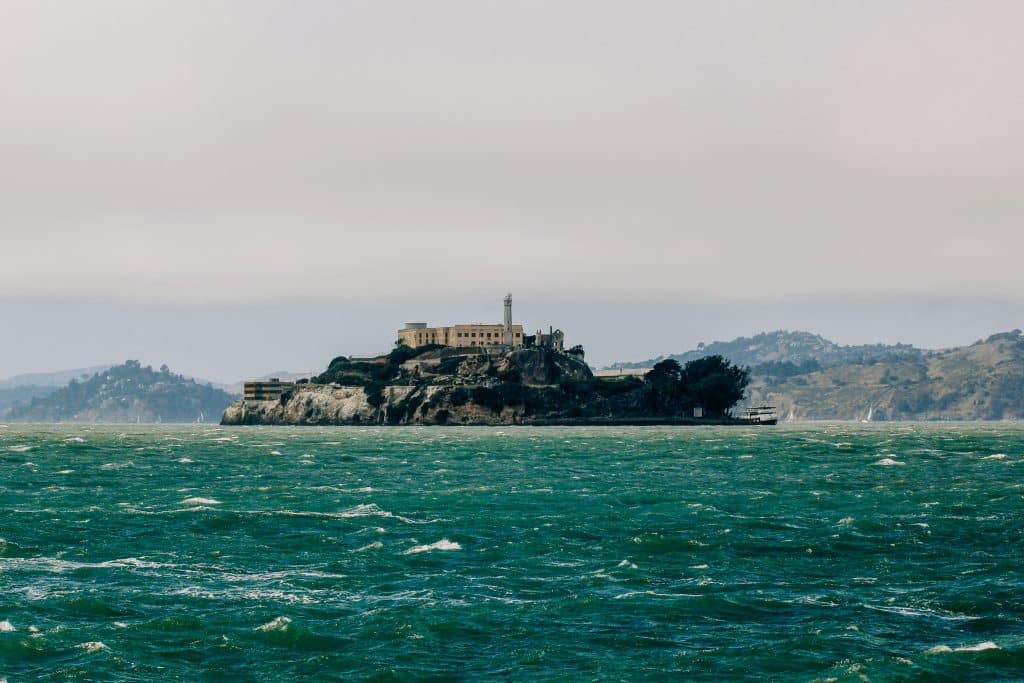 View of Alcatraz Island from the water, showing the historic prison buildings atop the rocky island, surrounded by the San Francisco Bay under a clear sky.