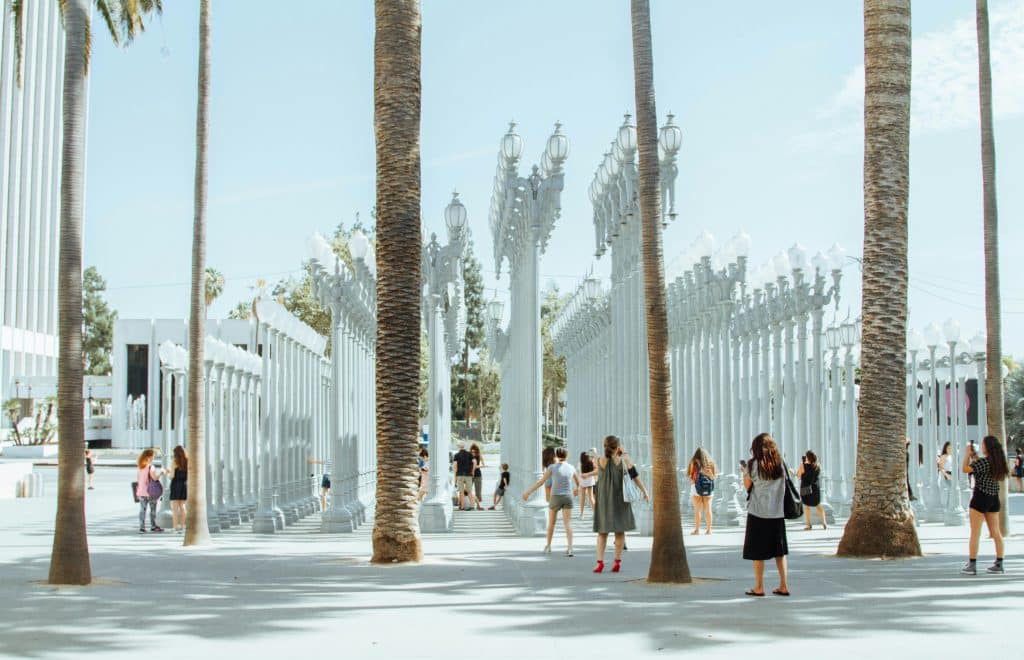 Installation of tall white cylindrical light poles outside LACMA (Los Angeles County Museum of Art), arranged in a grid on the plaza with the museum buildings in the background under a clear sky.