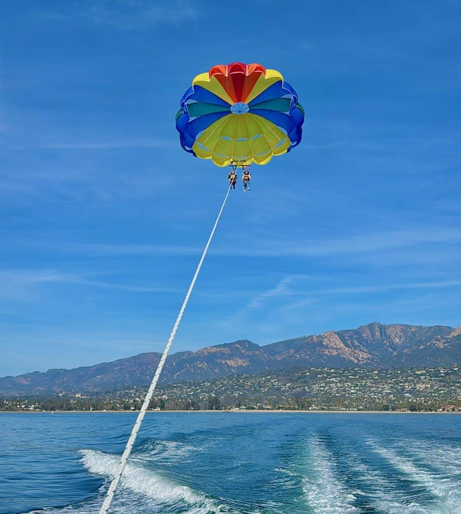 Mother and daughter parasailing over Santa Barbara Harbor, holding hands as they soar above the water, with coastal views of the city and shoreline in the background.