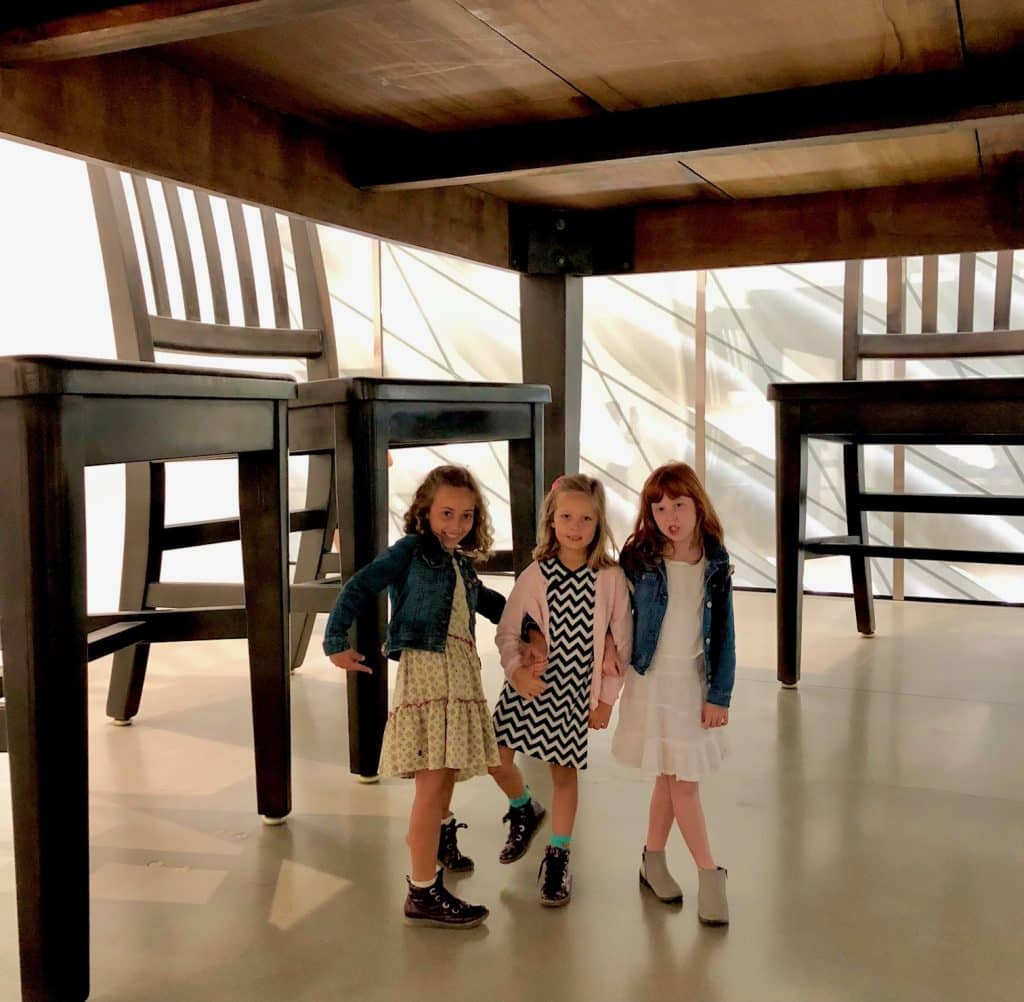 Three girls standing beneath a giant table installation at The Broad Museum, creating a playful forced-perspective illusion that makes them appear tiny