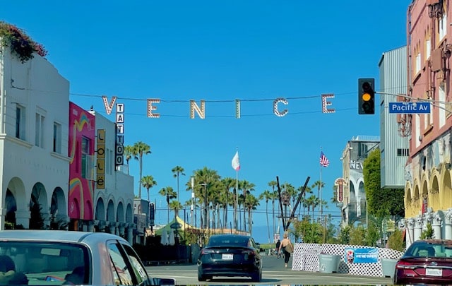 The iconic Venice Beach sign hanging above the street, reading ‘VENICE,’ with palm trees, storefronts, and blue sky in the background.