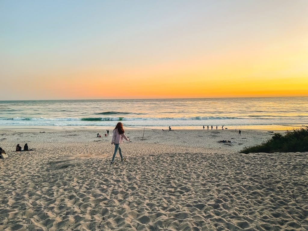 Girl walking along the sandy beach in Carmel, California, with the sun setting over the ocean, casting warm golden light across the shoreline.