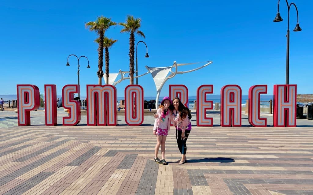 Mother and daughter standing together in front of the iconic Pismo Beach sign, smiling with the ocean and palm trees in the background.