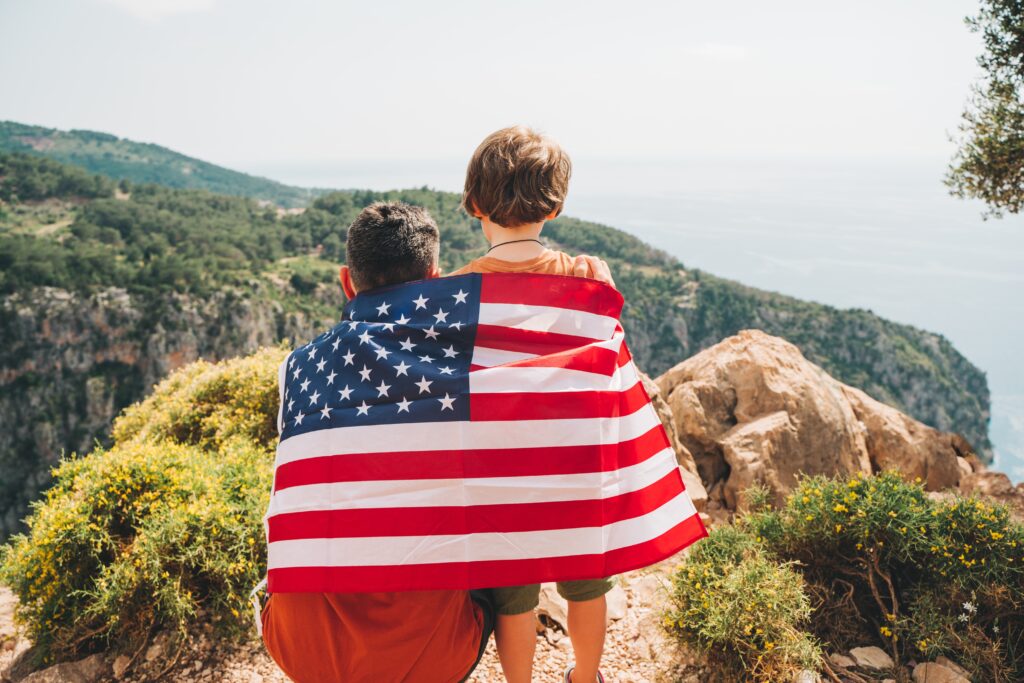 father and son wrapped in american flag sitting on mountain edge