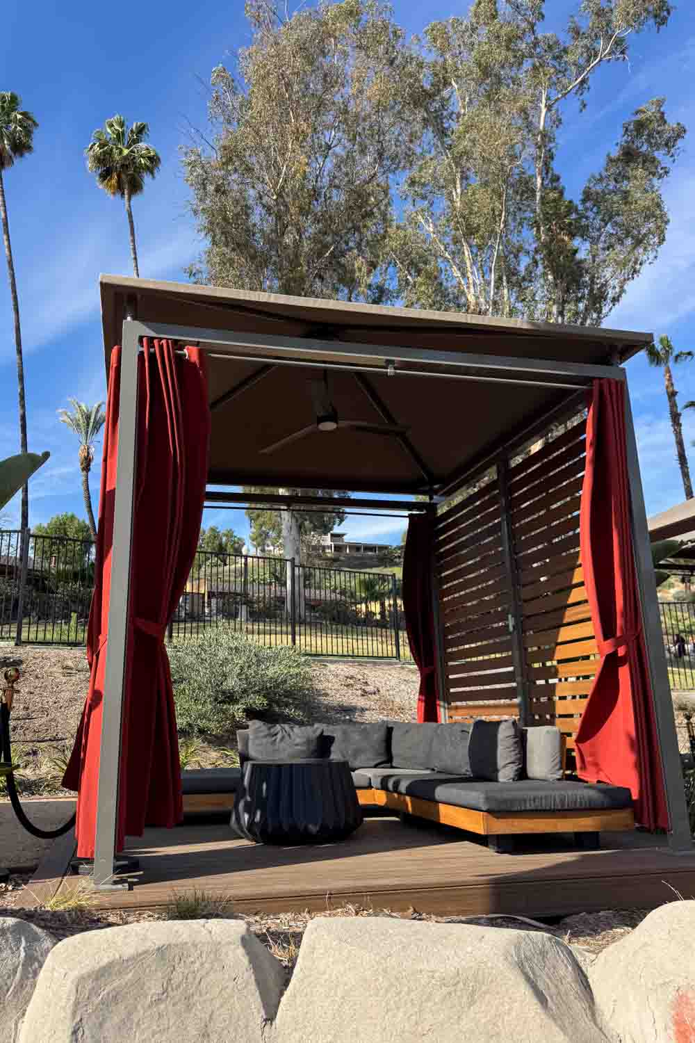 Outdoor resort cabana with red curtains. A gray cushioned sectional. And a black table under a shaded roof.