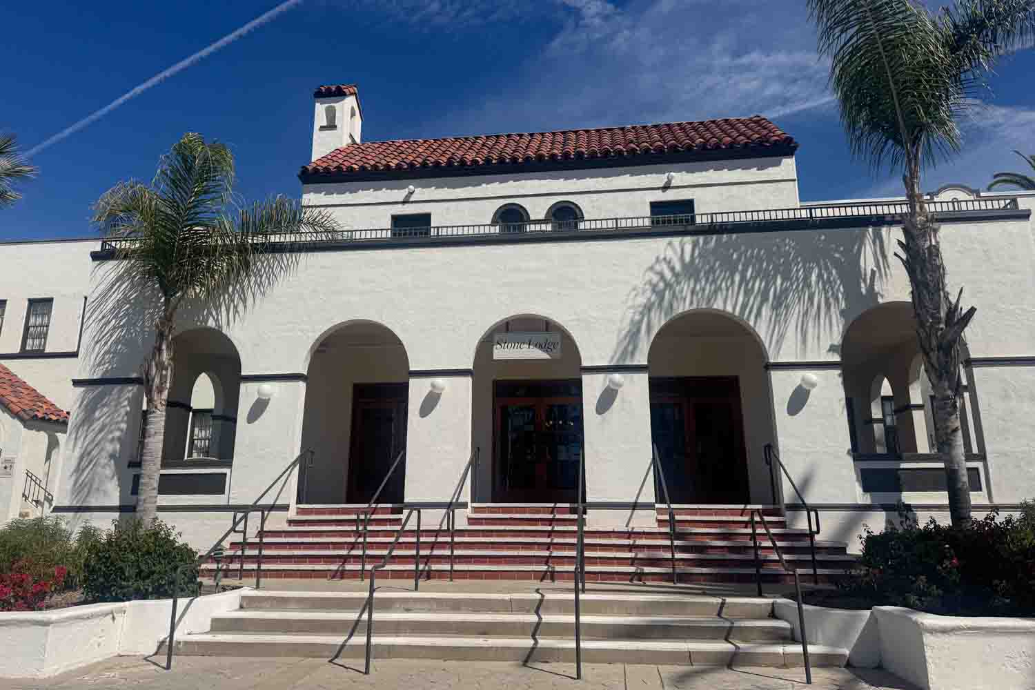 White Spanish style resort building with red tile roofing and arched entrances under a bright blue sky. The sign above the center entrance reads “Stone Lodge.”
