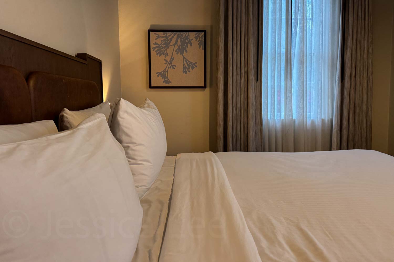 A hotel room bed with white pillows and bedding sits beside a curtained window. Warm lighting and simple wall art give the room a calm feel.