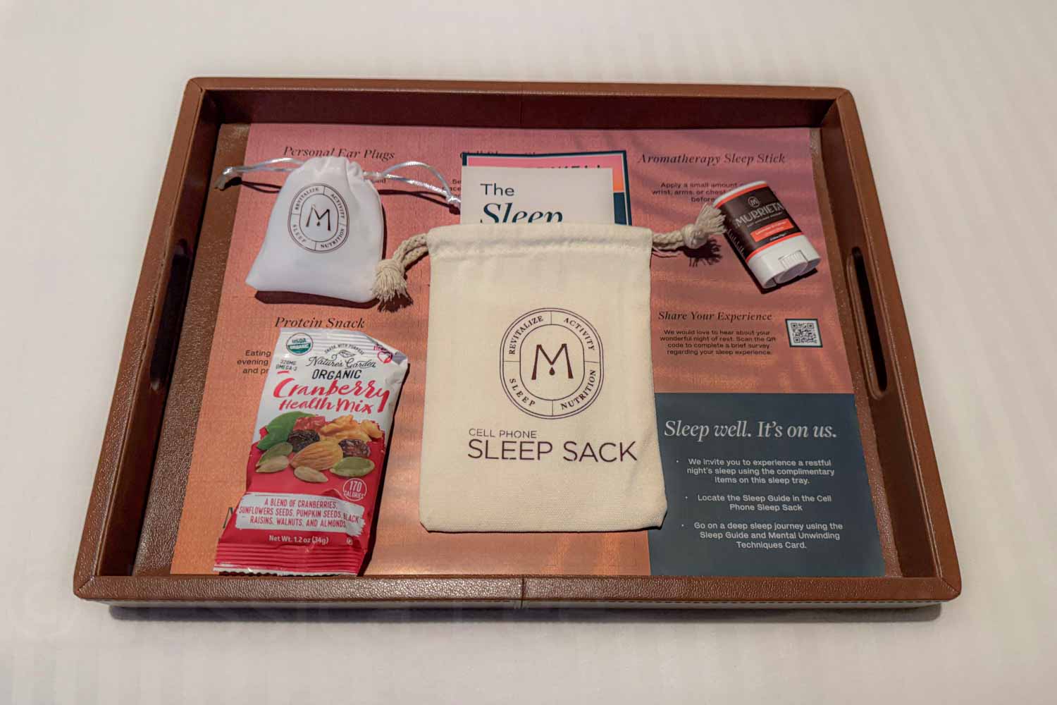 A wooden tray holds a cell phone sleep sack. Ear plugs. A protein snack. And a sleep guide card. The printed materials include the text “Cell Phone Sleep Sack” and “Sleep well. It’s on us.”