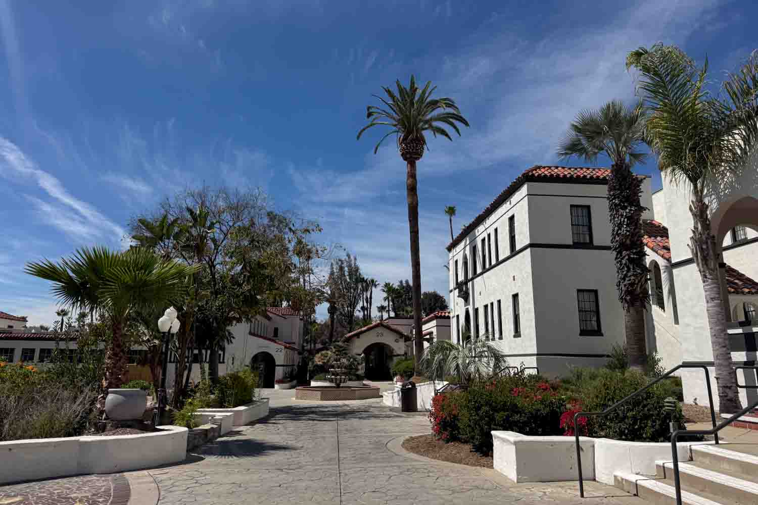 White Spanish style resort buildings with red tile roofs line a sunny courtyard. Palm trees and garden beds frame the walkway.