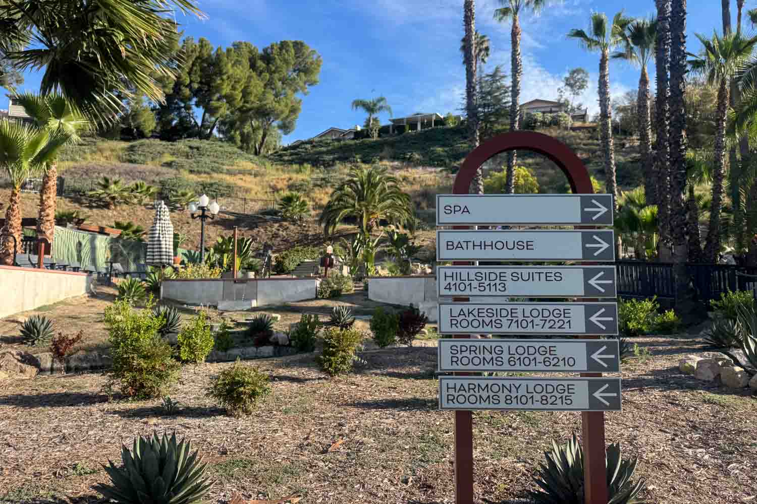 Resort directional sign in a landscaped desert garden with palm trees and a blue sky. The sign points to “SPA” “BATH HOUSE” “HILLSIDE SUITES 4101-5113” “LAKESIDE LODGE ROOMS 7101-7221” “SPRING LODGE ROOMS 6101-6210” and “HARMONY LODGE ROOMS 8101-8215”.