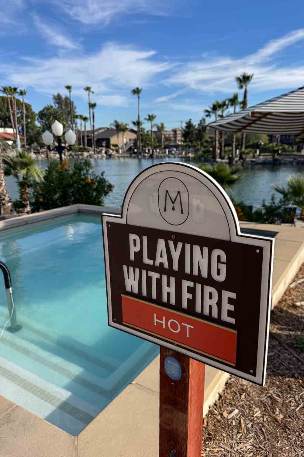 A sign beside a small pool reads “PLAYING WITH FIRE” and “HOT.” The lake and palm filled resort grounds are blurred in the background.