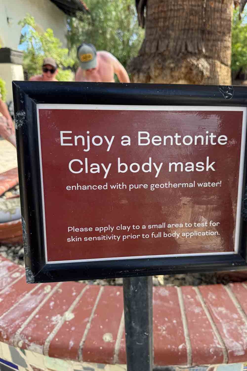 A sign beside a brick ledge reads “Enjoy a Bentonite Clay body mask enhanced with pure geothermal water. Please apply clay to a small area to test for skin sensitivity prior to full body application.” People stand blurred in the background near a palm tree.