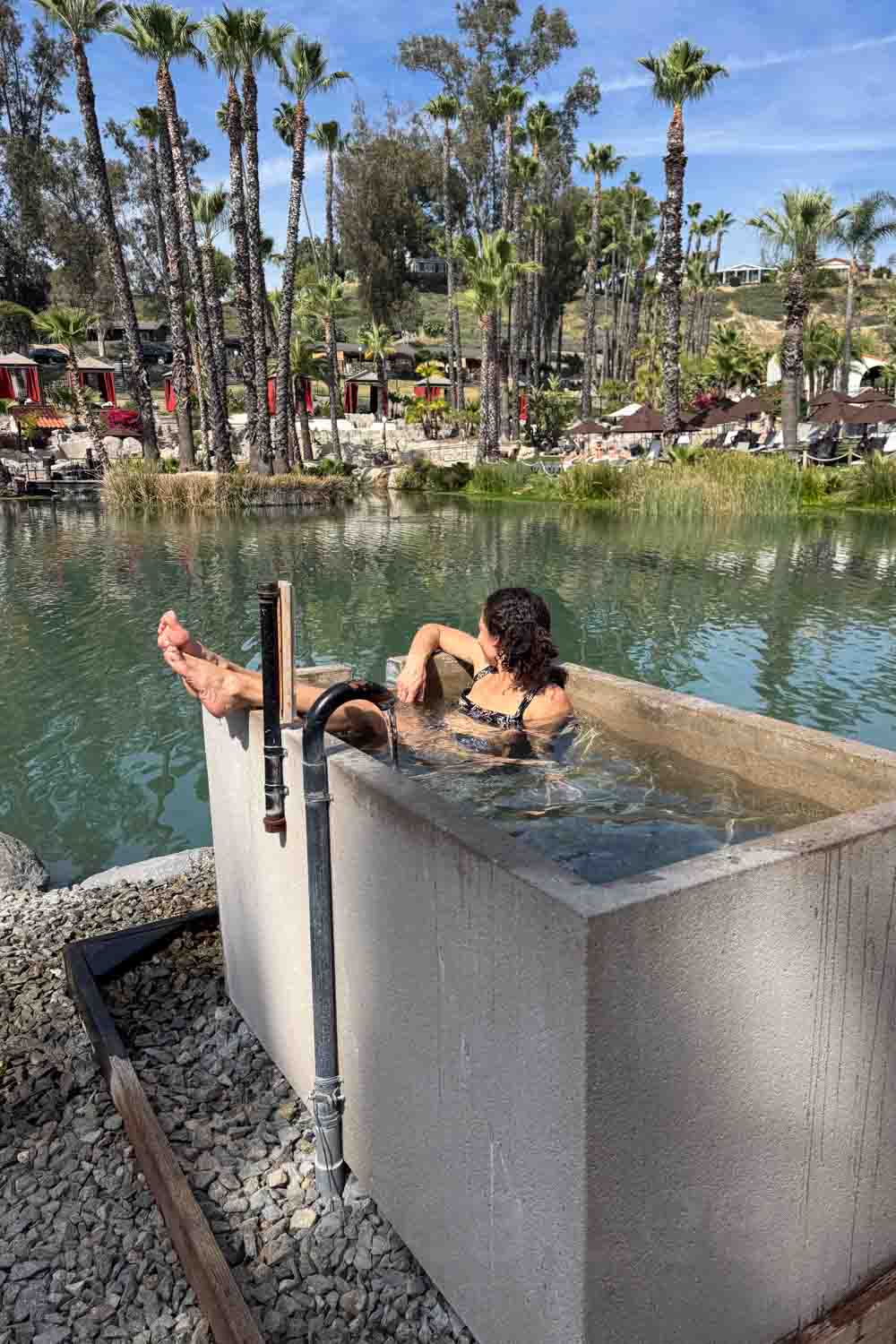 A woman relaxes in a rectangular outdoor hot tub beside a lake. Palm trees and resort cabanas line the opposite shore.