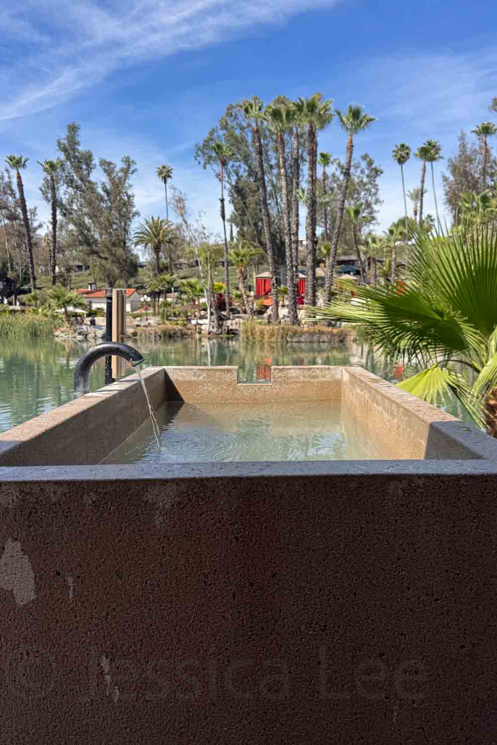 A concrete hot tub with a black water spout faces a palm lined lake. The view looks out toward resort landscaping and red cabanas across the water.