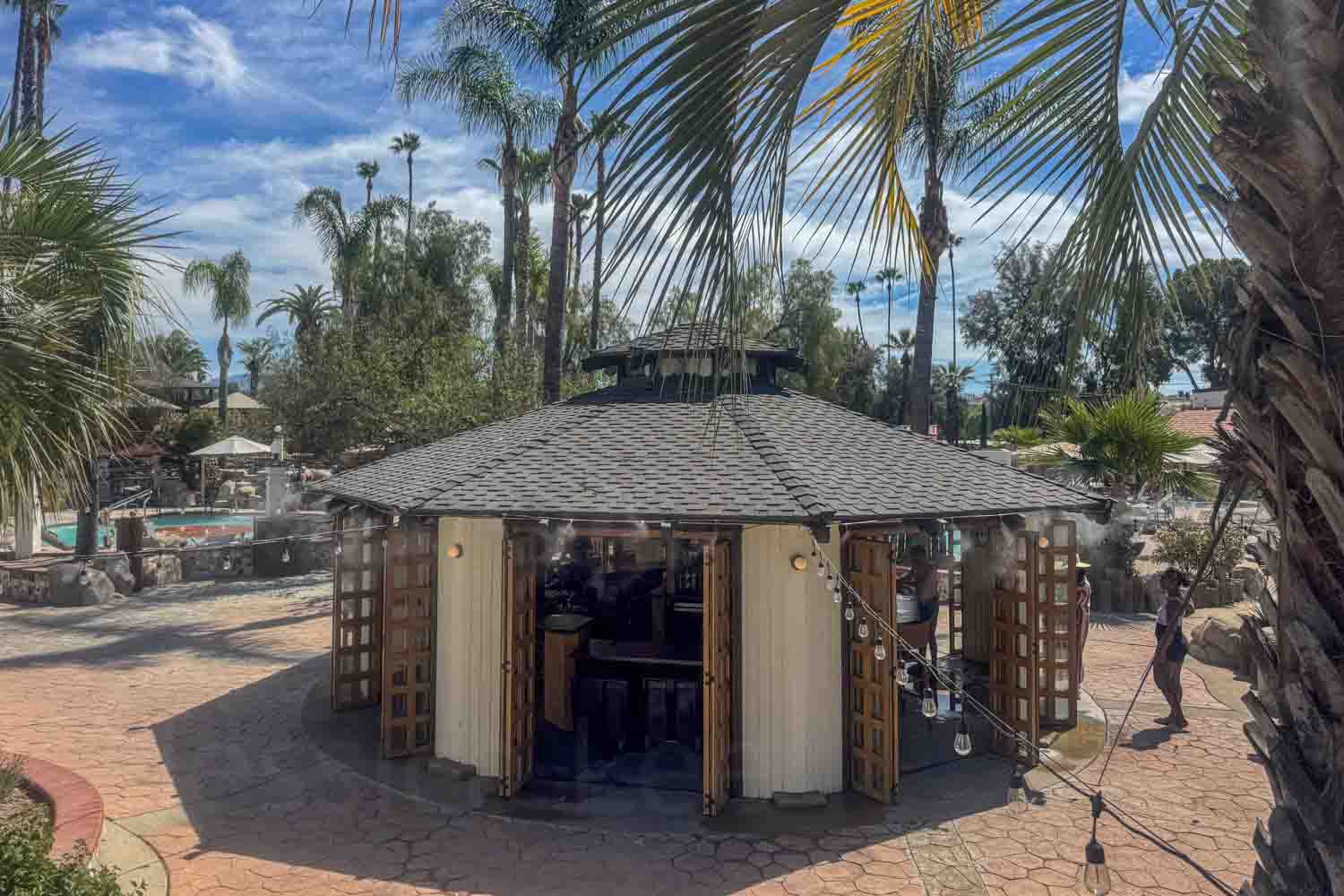 Palm trees frame a small pavilion with open wooden doors at a sunny resort courtyard. A pool and lounge area sit in the background.