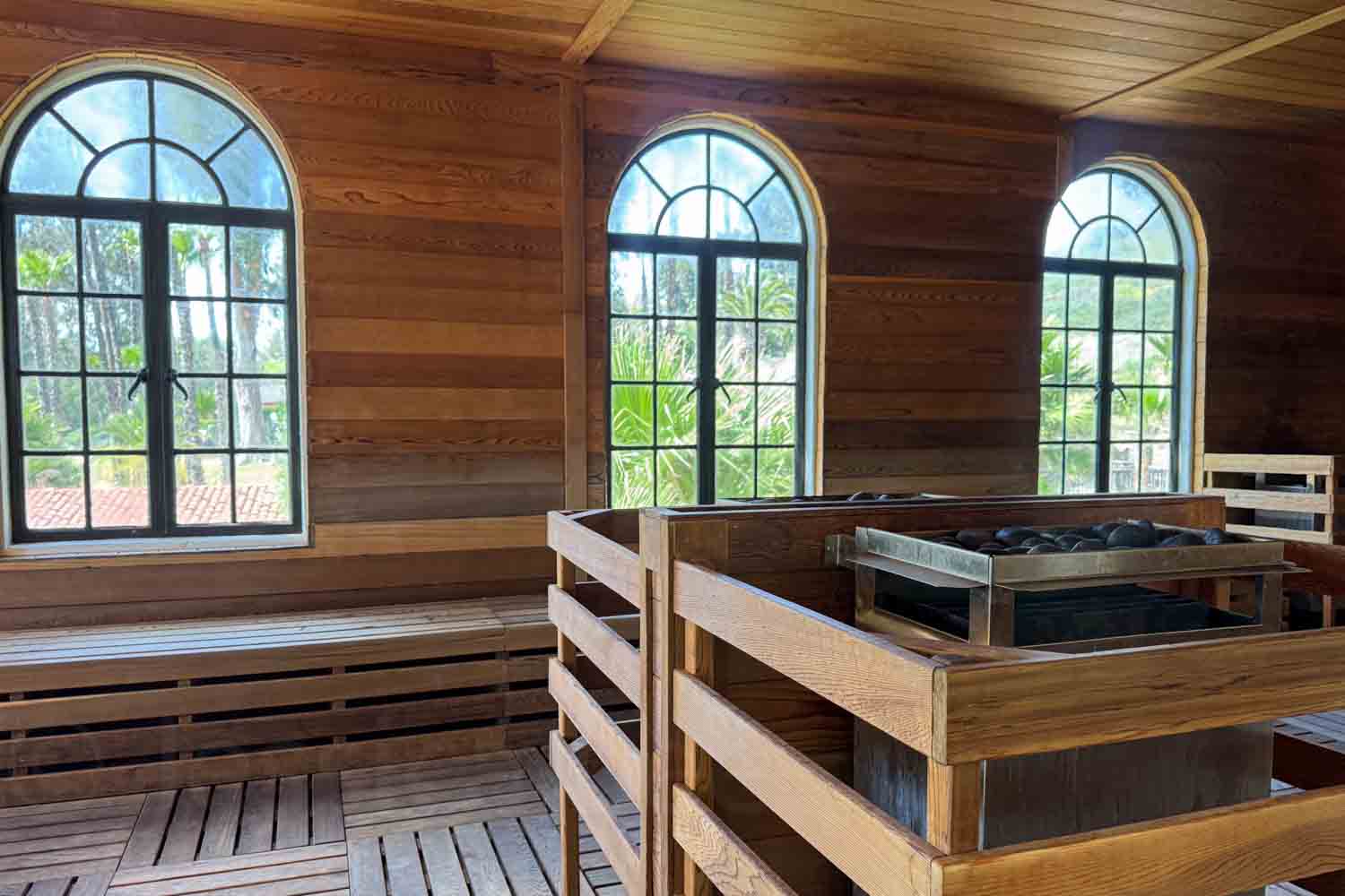 Wood-lined sauna room with arched windows. Long benches. And a central heater surrounded by a wooden guard rail.